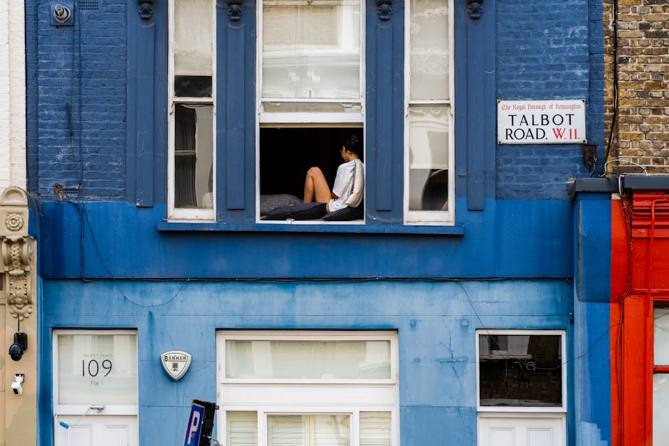 A young woman with dark hair sits on an open window ledge of a blue-painted residential building in Kensington, with her legs hanging outside and her back resting against the window frame. She is wearing a white top and appears to be looking down or lost in thought. The window is open, revealing a glimpse of an interior room behind her, and the exterior features wooden window trim and shutters. To the left of the window is a partially visible facade of adjacent buildings, with a street sign reading 'Talbot Road, W.11' attached to the building's brick wall. Below the window, a white door numbered '109' with a small window and a building security alarm are visible. The property is part of a typical London house, with a front facade painted in a vibrant blue, contrasting with surrounding buildings' brickwork. This scene may illustrate home relocation or moving preparations, possibly involving packing and loading furniture or boxes from the upper floor, with the presence of a residential environment associated with house removal services. The overall setting suggests a quiet urban street in Kensington, captured during daylight.