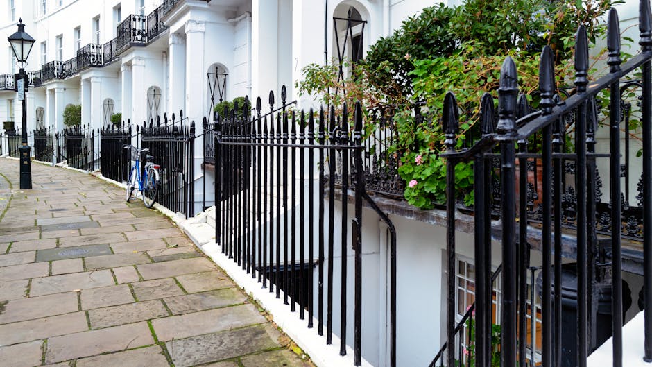 A row of white terraced houses with black wrought iron front fences lining a paved sidewalk in Kensington, London. The fences feature pointed finials and are positioned along the edge of the buildings, separating the footpath from the residential properties. One house has a small set of stairs leading up to its entrance, partially obscured by greenery and flowering plants in pots placed on the front steps. A blue bicycle is leaning against the fence near the sidewalk. The scene is illuminated by natural daylight, with lush foliage visible above the fences, suggesting an urban setting ideal for house removals and home relocation services. Man and a Van Kensington occasionally operate in such areas, assisting with furniture transport and packing as part of their removals service.