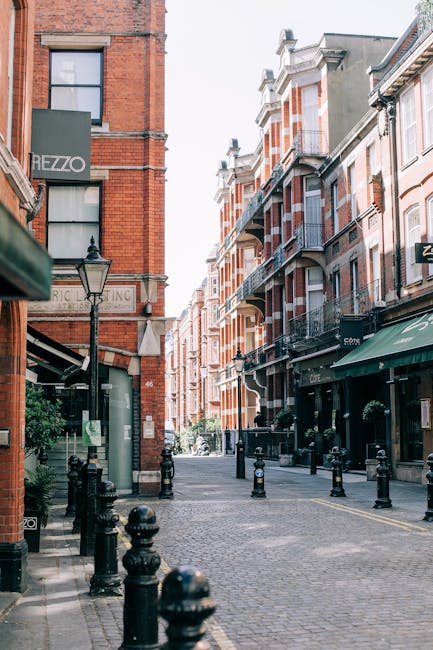 A narrow cobblestone street in a city area during daytime, lined with multi-storey brick and stone buildings featuring bay windows and balconies. On the left, a tall red-brick building with a sign reading 'HEZZO' is visible, along with a traditional-style lamppost positioned on the pavement. To the right, shops with green awnings and glass windows are present, while black bollards line the edges of the road to control vehicle access. The street appears quiet with no visible pedestrians or moving vehicles. This setting illustrates typical urban surroundings where household removals, such as packing and furniture transport, may take place, with the alleyway serving as a potential loading or unloading area for a professional moving service like Man and a Van Kensington, involved in home relocation and furniture moving operations.