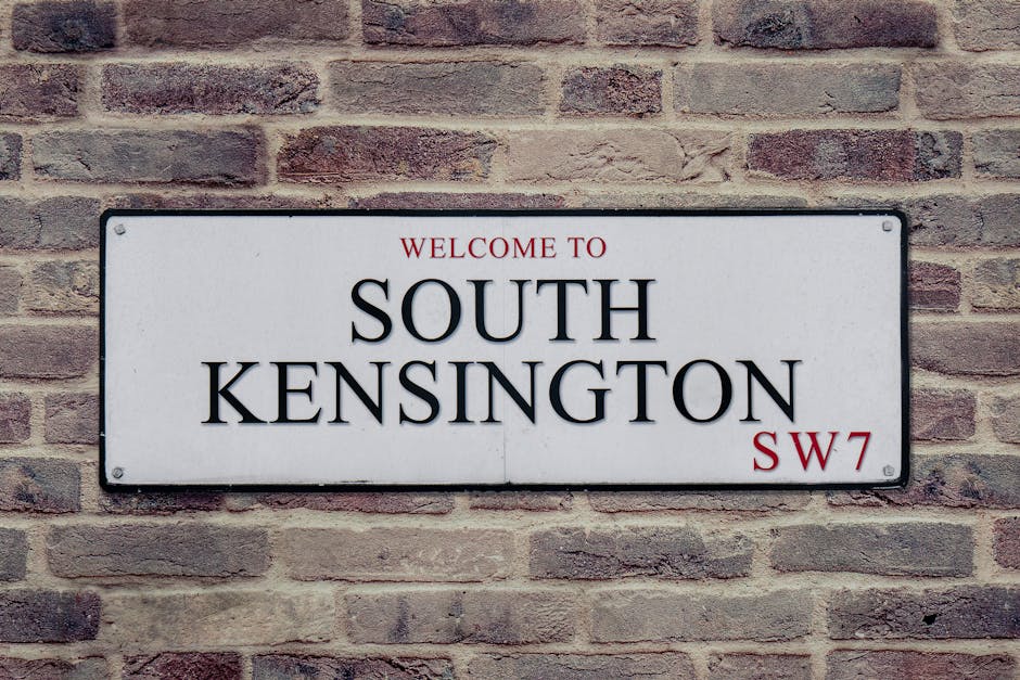 A rectangular white sign with black and red text is mounted on a brick wall, displaying 'Welcome to South Kensington SW7'. The sign is attached with four metal bolts, one in each corner. The wall behind the sign consists of reddish-brown bricks arranged in a running bond pattern with visible mortar joints. The lighting appears natural, illuminating the sign evenly, and the bricks have a textured surface. This signage is relevant to house removals and moving services in the South Kensington area, where [COMPANY_NAME] may assist with furniture transport and packing during a home relocation process.