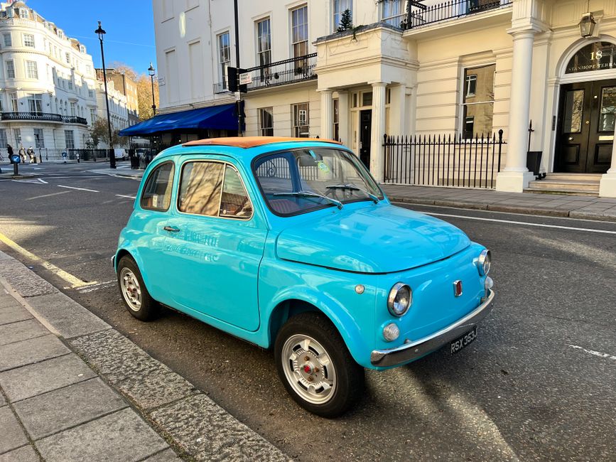 A small vintage blue car parked on a city street in front of a white building with black railings and an address plaque. The car has a rounded, compact shape typical of classic European city vehicles, with a tan fabric roof, circular headlights, and small steel wheels. It is situated close to the curb with a parking lane marked by a yellow line, and the pavement is partly shaded by nearby buildings. In the background, there are additional parked cars, pedestrians, and typical urban street furniture. The scene is lit by daylight, indicating clear weather. This setting illustrates the type of vehicle associated with home relocation or furniture transport services provided by Man and a Van Kensington, often used for local removals or moving logistics within urban areas such as the Royal Borough of Kensington and Chelsea.