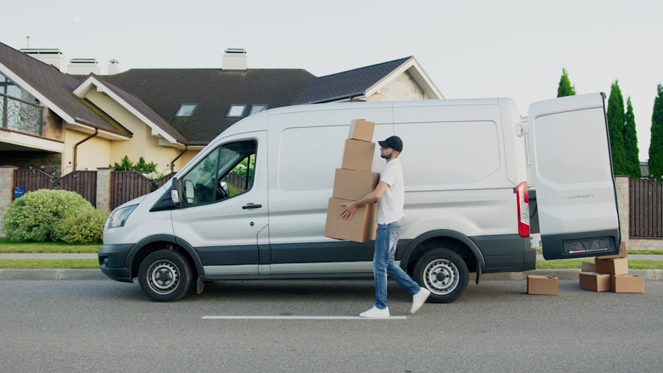 A man wearing casual clothes, including a white t-shirt, blue jeans, and a black cap, is carrying three stacked cardboard boxes during a home relocation, standing beside a white cargo van with its side door open. The van is parked on a residential street in front of a modern, detached house with beige and brown exterior walls, a dark roof, and large windows. Inside the van’s open compartment, additional cardboard boxes and packing materials are visible, indicating a packing and moving process. The sidewalk is lined with greenery, including bushes and tall conifer trees, and the scene is lit by daylight, showcasing an organized furniture transport operation by Man and a Van Kensington for domestic removal services, consistent with the page about same day house removals and delays in Kensington.