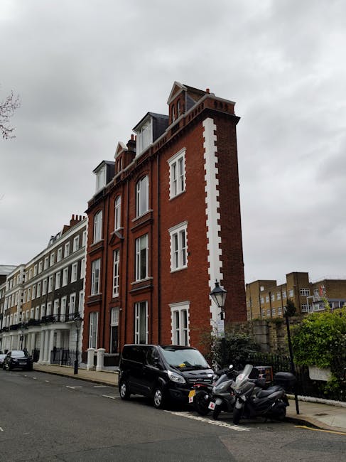 A multi-storey residential building with a red brick facade and white decorative elements, situated on a street in Kensington under an overcast sky. The building features large sash windows and a small attic dormer at the top. In the foreground, a black van is parked alongside a group of motorcycles and scooters on the pavement, indicating a busy urban environment. The street is lined with similar Victorian-style houses, some painted in muted colours, with a row of parked cars and a lamppost visible nearby. The exterior surroundings suggest a typical London neighbourhood, suitable for home relocations and furniture transport services provided by Man and a Van Kensington, as part of their removals and packing operations.