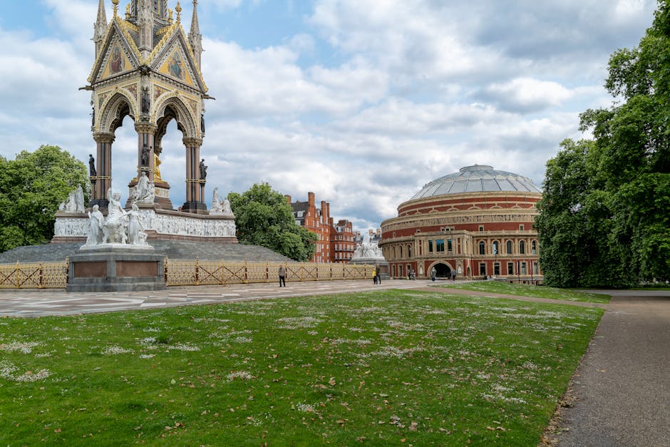 The image shows a large, ornate monument with multiple statues and decorative elements, situated on a paved area in a park. The monument features a tall, intricate structure with a pointed spire at the top, and is surrounded by low fencing. In the background, there is a historic building with a rounded, domed roof and a red brick exterior, alongside lush green trees on either side. The sky above is partly cloudy, with patches of blue visible through the clouds. The park area is well-maintained, with a grassy lawn bordered by a pathway. The scene appears to be a daylight setting with natural light illuminating the monument, building, and surrounding greenery. Occasionally, individuals can be seen walking in the distance. This setting illustrates a public space that may be involved in various events, including those related to moving or setting up portable items, though no specific activity is visible in this moment. Man and a Van Kensington specializes in house removals and furniture transport, and this image captures a typical urban park environment where such services could operate to facilitate home relocations in the W8 postcode area, including parking and logistics considerations for moving companies.