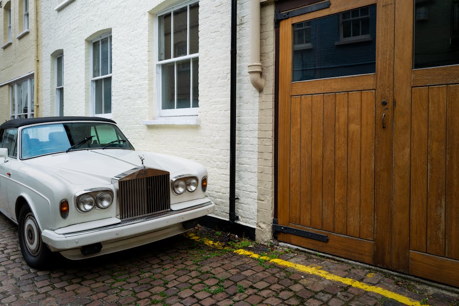 A vintage white car with a black soft top parked on a cobbled driveway next to a cream-colored brick house with white-framed sash windows. The car's front grille and round headlights are clearly visible, and it is positioned close to a large wooden garage door with black hardware. Inside the garage, partially visible are packed cardboard boxes wrapped in plastic and brown paper, alongside a large wooden packing crate and a clothing item in fabric covering. The scene is outdoors during daytime, with natural light illuminating the area, and a black downspout running alongside the house. This setting depicts a home relocation scenario, with furniture and packaging materials prepared for transport, highlighting the process of loading and packing items during a move, as managed by [COMPANY_NAME], operating in the area of removals and house moves for the W8 postcode with appropriate parking arrangements.