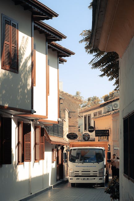 The image shows a narrow street lined with light-colored buildings featuring wooden shutters and small balconies, with a white van parked on the cobbled pavement in the center. The van is positioned between two buildings, with its front facing the camera and partially obstructed by the surrounding structures. The street appears to be in a residential or commercial area, with signs visible on some of the buildings and a person walking nearby on the right side. The scene is illuminated by natural daylight, and some greenery from trees is visible overhanging the rooftops. This setting reflects a typical environment encountered during home relocation or furniture transport in restricted access conditions, which [COMPANY_NAME], as suggested by the context on the provided webpage, might work to navigate efficiently during moving and packing processes. The image captures the logistical challenge and environment typical for professional removals services involved in house removals or furniture transport within tight urban spaces.