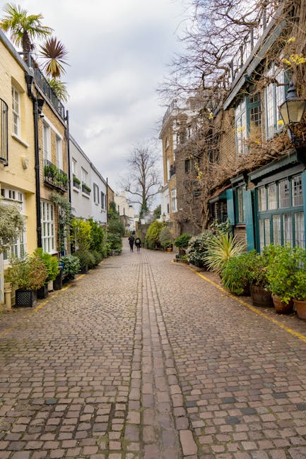 A cobbled residential street in Kensington featuring narrow access with closely spaced, multi-storey houses on both sides. The houses have a variety of exterior styles, with some painted in light colours and others with brick or timber cladding, and many are adorned with potted plants and small garden areas. The street is lined with potted shrubs and small trees, adding greenery along the pavement. In the distance, two people are walking, and the sky is overcast with grey clouds. The scene demonstrates typical urban surroundings where a relocations specialist such as Man and a Van Kensington may need to address narrow access challenges during home removals, involving careful furniture transport and loading processes through limited space.